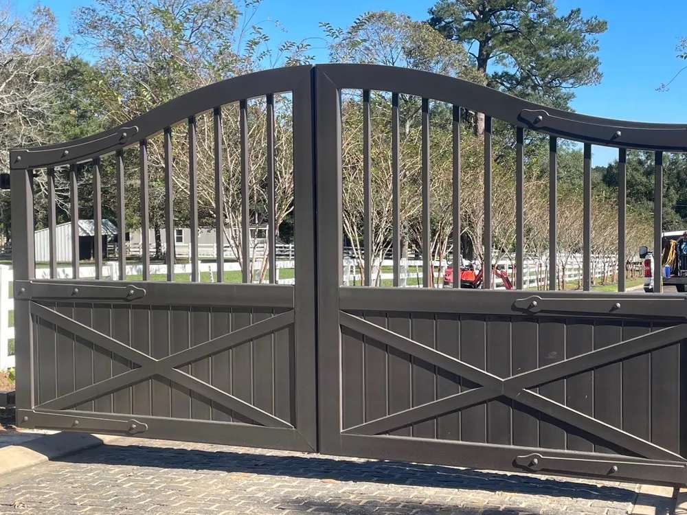 The Stable Gate closed across a paver drive, its arched bronze leaves boarded and X-braced below open pickets, with white paddock fencing and a barn behind.