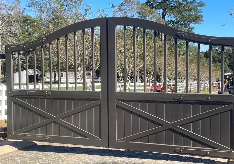 The Stable Gate closed across a paver drive, its arched bronze leaves boarded and X-braced below open pickets, with white paddock fencing and a barn behind.