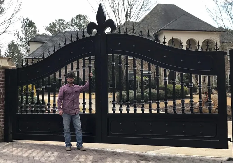 The Scout Gate closed across a paved drive, its curve rising to a fleur-de-lis at the centre, with a man standing against the left leaf for scale and a large brick and stone house behind.