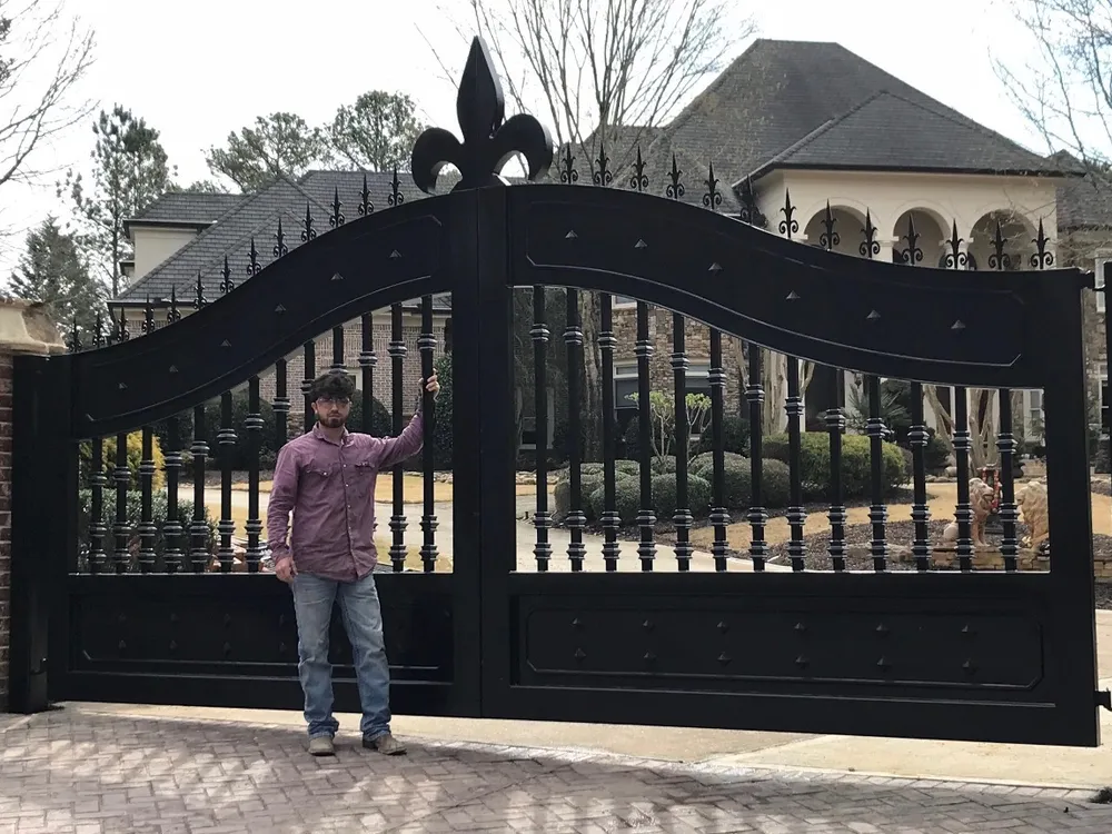 The Scout Gate closed across a paved drive, its curve rising to a fleur-de-lis at the centre, with a man standing against the left leaf for scale and a large brick and stone house behind.