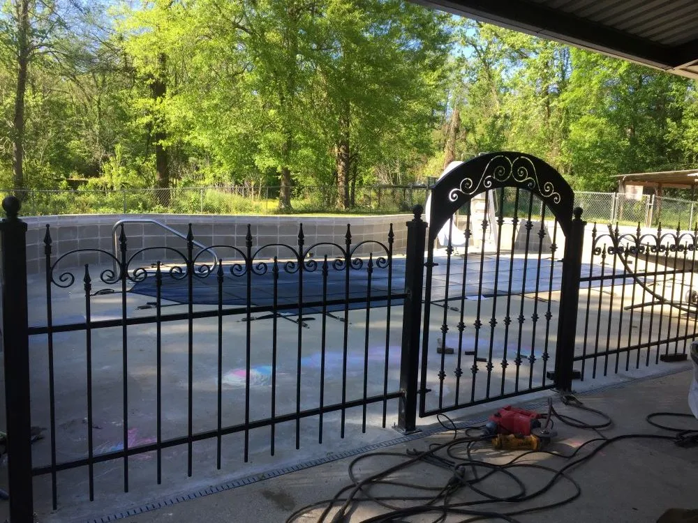 A black iron pool fence with an arched gate enclosing a swimming pool, wood privacy fencing and trees beyond.