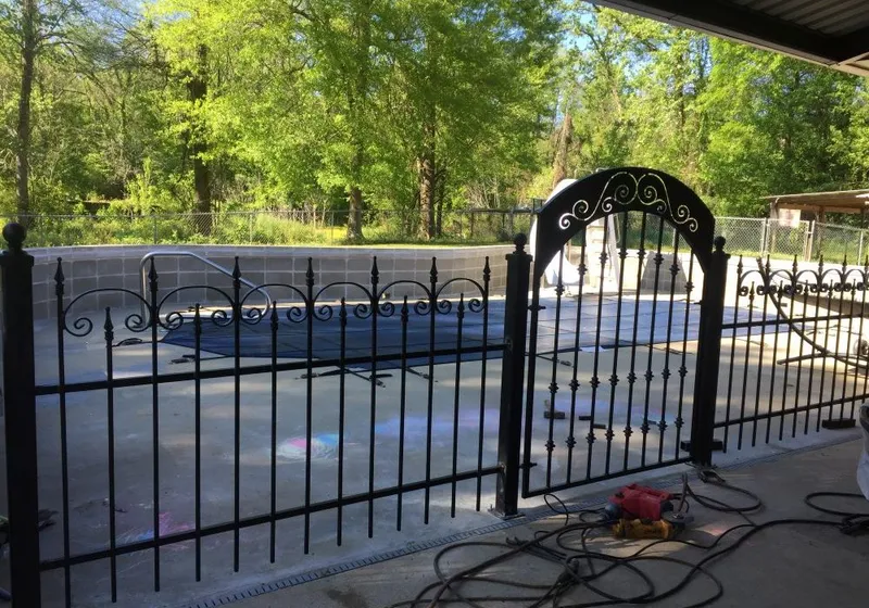 A black iron pool fence with an arched gate enclosing a swimming pool, wood privacy fencing and trees beyond.