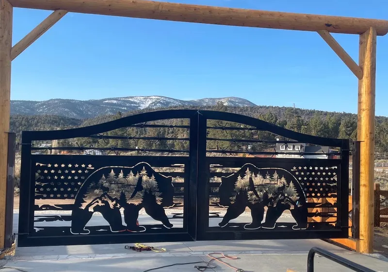 The Frontier Gate closed under a peeled-log header on a new concrete pad, bears and mountain peaks cut through its flag panel, with scrub and snow-dusted mountains beyond.