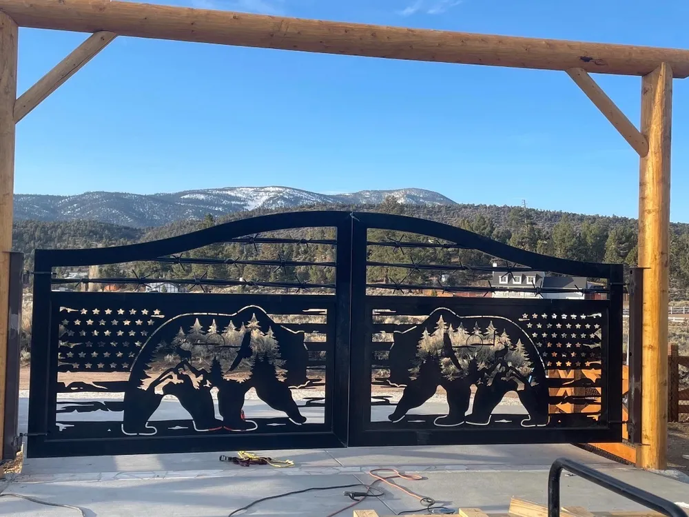 The Frontier Gate closed under a peeled-log header on a new concrete pad, bears and mountain peaks cut through its flag panel, with scrub and snow-dusted mountains beyond.