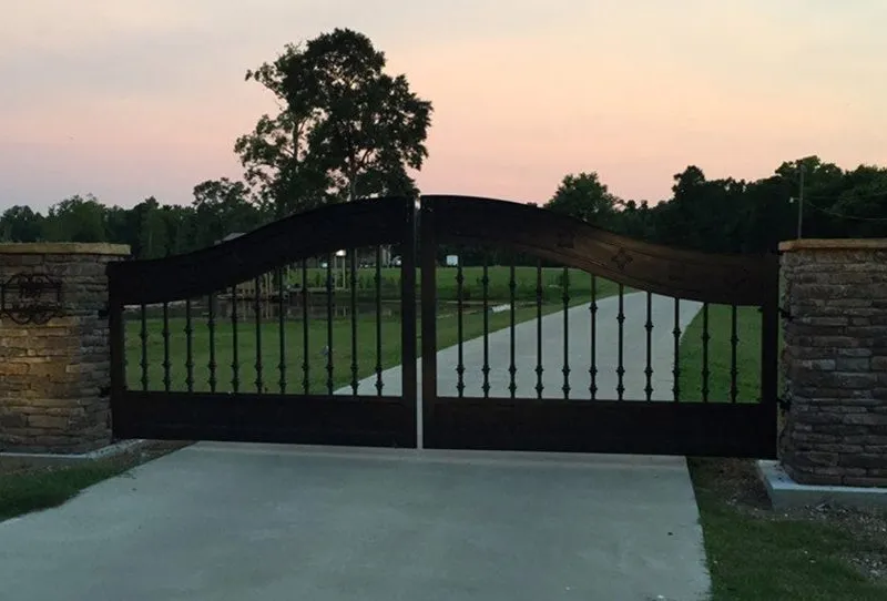 The Ham Gate closed across a concrete driveway at dusk, its curved iron top rail silhouetted against an orange sunset sky between stone columns.