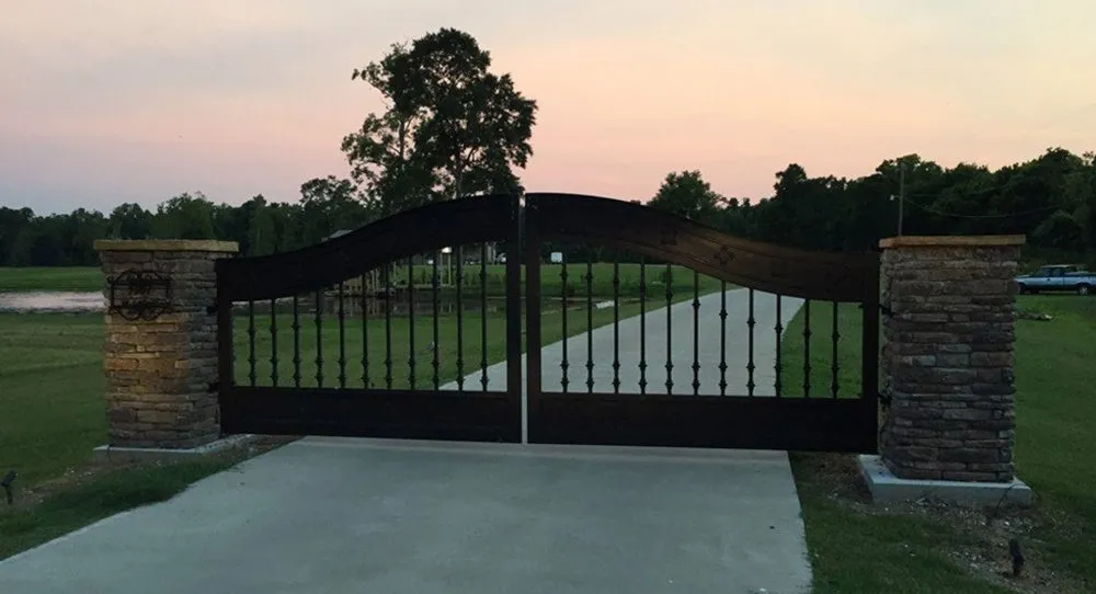 The Ham Gate closed across a concrete driveway at dusk, its curved iron top rail silhouetted against an orange sunset sky between stone columns.