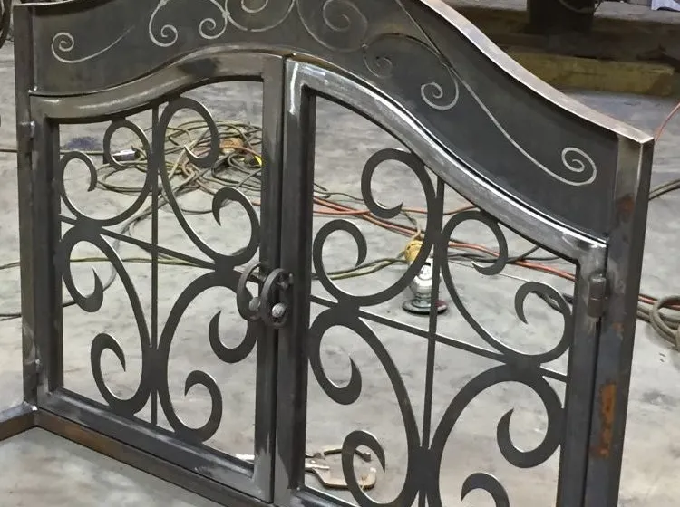 A double-leaf arched courtyard gate in raw burnished steel, both leaves filled with curling formed scrollwork and the arched header scribed with matching scroll lines, standing on the fabrication shop floor.