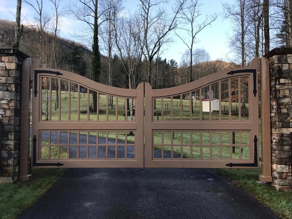 The Clay Gate closed across an asphalt drive between two stone columns, its brown grid leaves dipping to the centre with pasture, bare trees and a wooded ridge beyond.