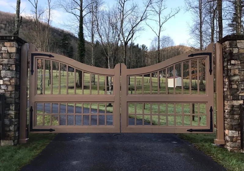 The Clay Gate closed across an asphalt drive between two stone columns, its brown grid leaves dipping to the centre with pasture, bare trees and a wooded ridge beyond.