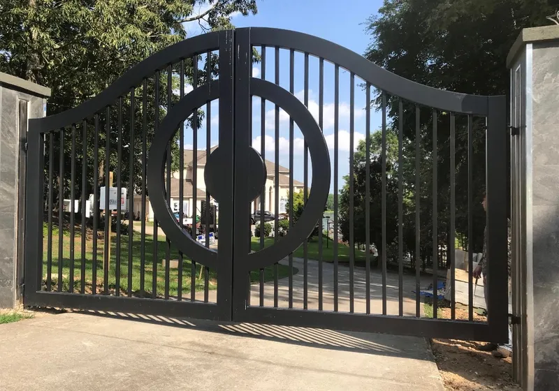 The Lake Spivey Gate closed square-on across a concrete drive, a split circle with a round centre boss straddling the joint between its two picket leaves, with lawn, trees and a pale house beyond and stone-clad pillars to either side.