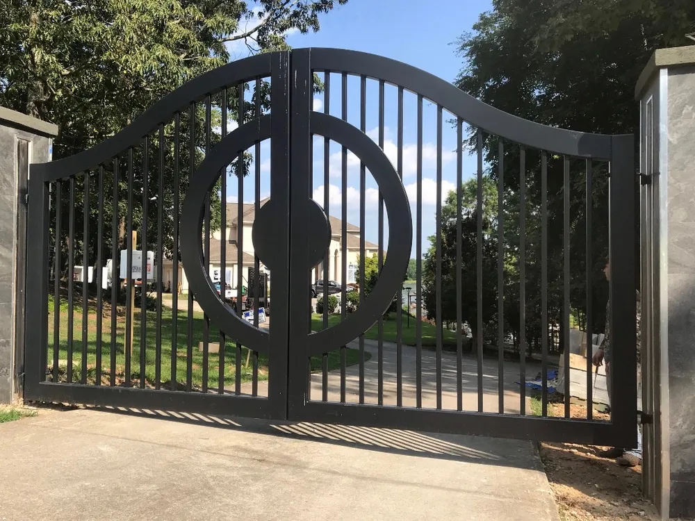 The Lake Spivey Gate closed square-on across a concrete drive, a split circle with a round centre boss straddling the joint between its two picket leaves, with lawn, trees and a pale house beyond and stone-clad pillars to either side.