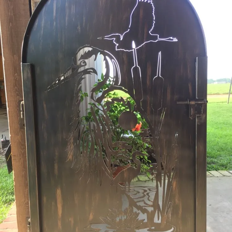 An arched steel walk gate with a marsh scene cut through the solid plate — a great blue heron among cattails, a heron flying above and a duck on the water — daylight and a flowering plant showing through the cutouts.