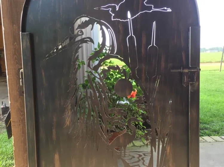 An arched steel walk gate with a marsh scene cut through the solid plate — a great blue heron among cattails, a heron flying above and a duck on the water — daylight and a flowering plant showing through the cutouts.