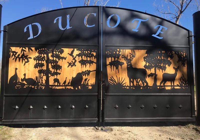 The Backcountry Gate closed across a dirt drive, a name cut through its arched top panel and a silhouetted scene of deer, turkeys and ducks pierced through the middle band, lit from behind by timber boarding.