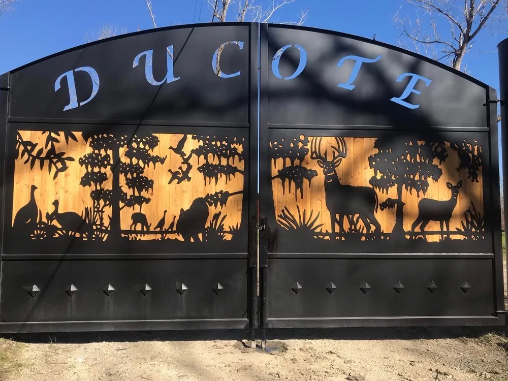 The Backcountry Gate closed across a dirt drive, a name cut through its arched top panel and a silhouetted scene of deer, turkeys and ducks pierced through the middle band, lit from behind by timber boarding.