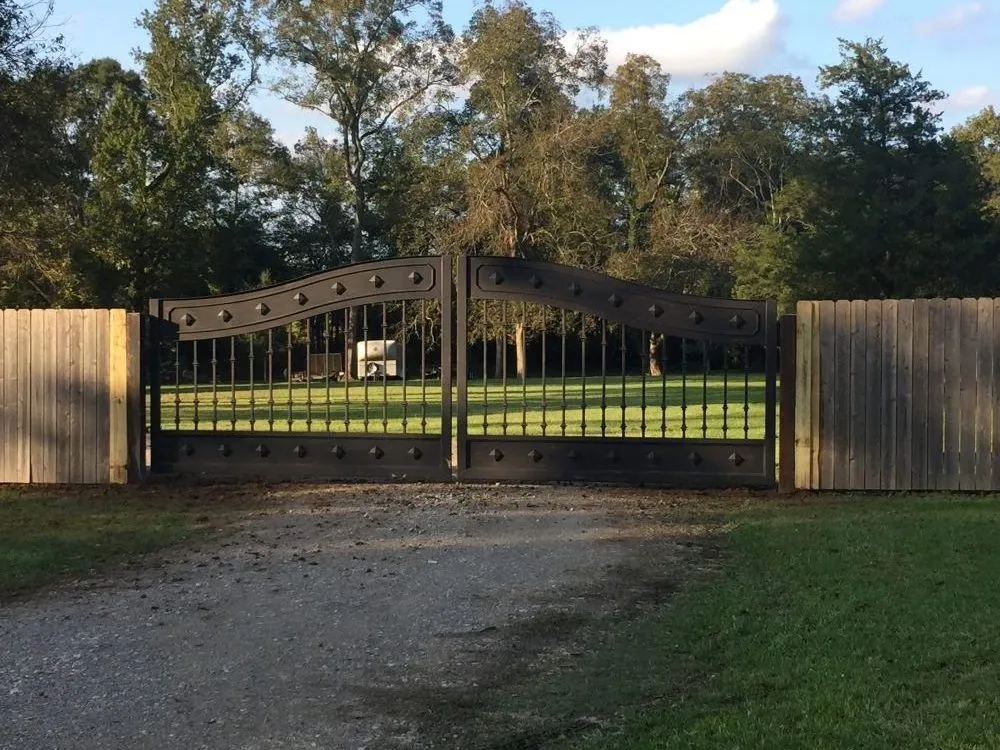 The Mayeaux Gate closed across a gravel driveway, its dark iron leaves set between wood privacy fencing with lawn and trees behind.
