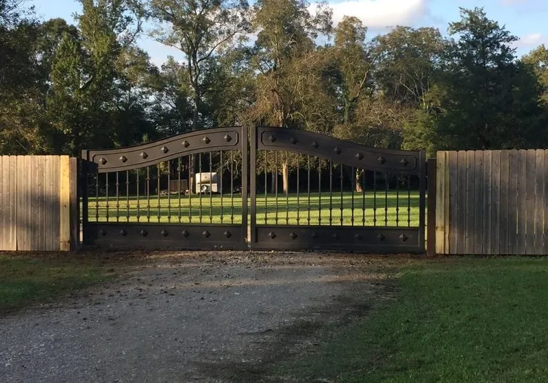 The Mayeaux Gate closed across a gravel driveway, its dark iron leaves set between wood privacy fencing with lawn and trees behind.