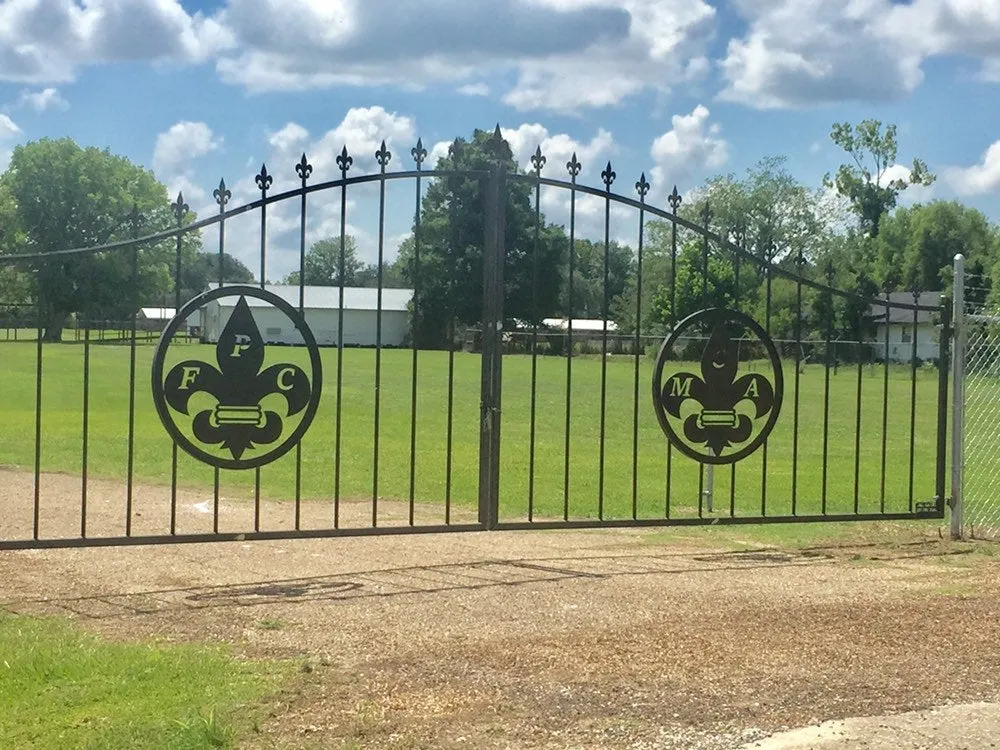 The MCA Gate closed across a drive, with fleur-de-lis finials along its arched top rail and a circular fleur-de-lis medallion set in each leaf.