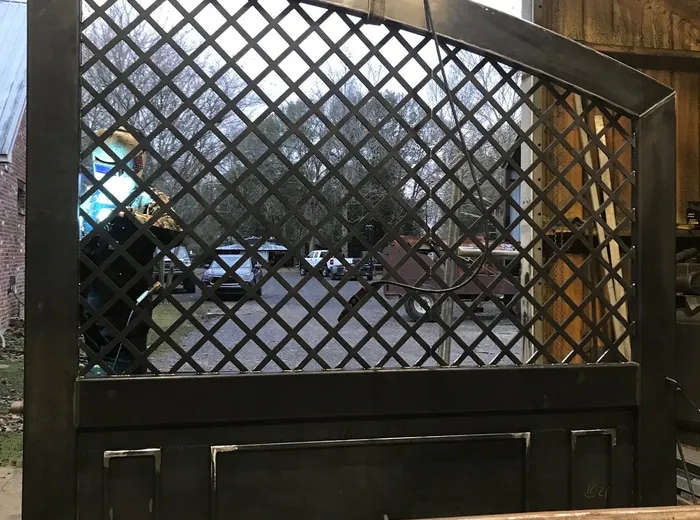 A single leaf hanging from a hoist in the shop before finishing, squared up on stands in bare steel, its diamond lattice of flat bar filling the upper frame above a solid panelled base.