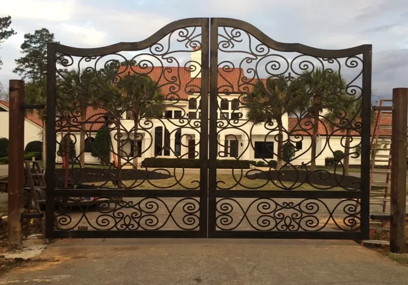 The Inman Gate closed across a drive, its scrollwork-filled leaves rising to a serpentine top rail, with a white tile-roofed house and palms visible through the ironwork.