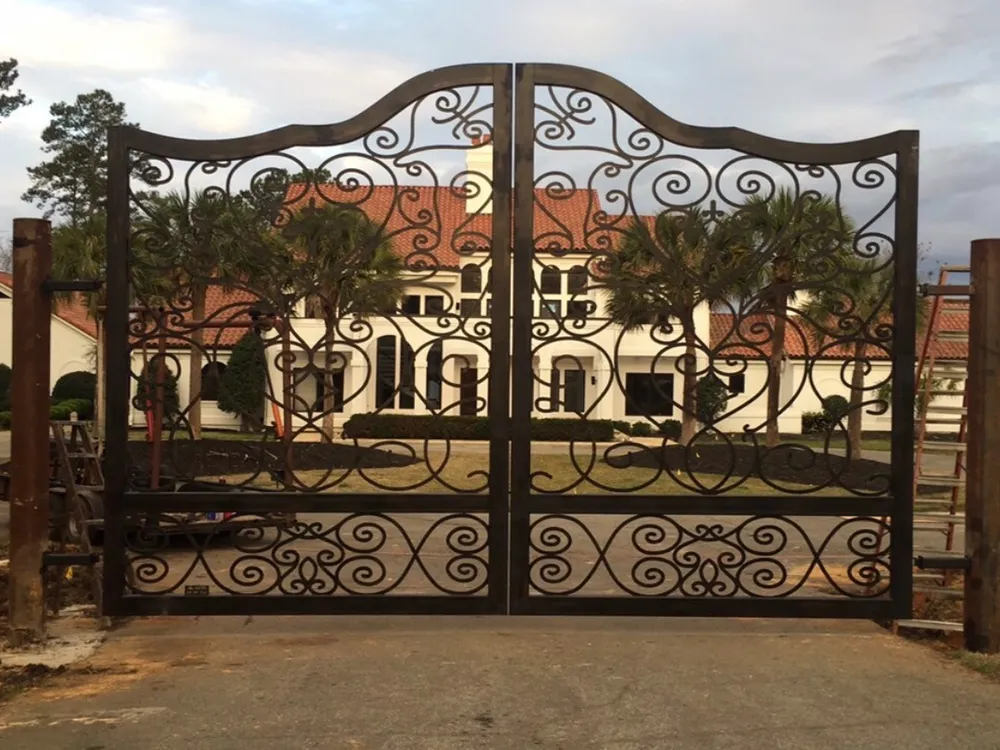 The Inman Gate closed across a drive, its scrollwork-filled leaves rising to a serpentine top rail, with a white tile-roofed house and palms visible through the ironwork.