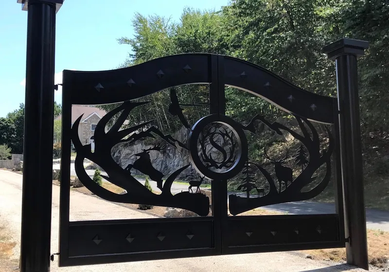 The Wilderness Gate closed between square black posts with stepped caps, its cut-steel scene of antlers, mountains, pines and deer in full sun, with a granite outcrop, trees and a stone-faced house visible through the openings.