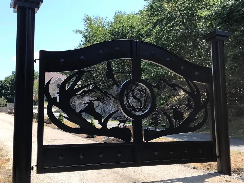 The Wilderness Gate closed between square black posts with stepped caps, its cut-steel scene of antlers, mountains, pines and deer in full sun, with a granite outcrop, trees and a stone-faced house visible through the openings.