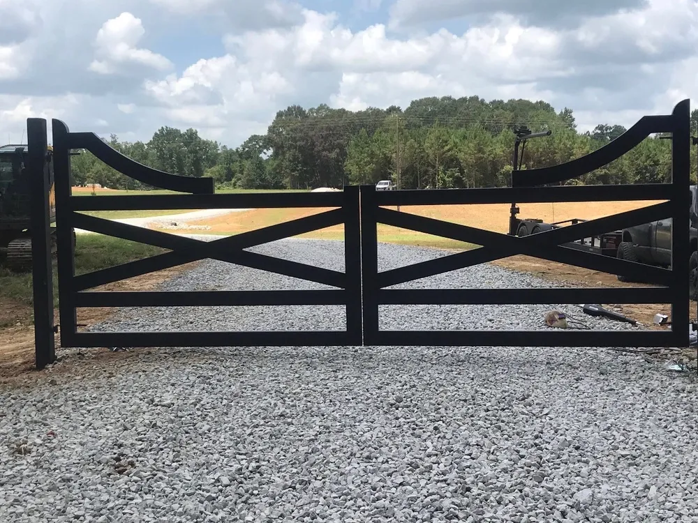 The Farmhouse Gate closed across a fresh gravel drive, two black X-braced leaves with scrolled top corners, open fields and a treeline behind.