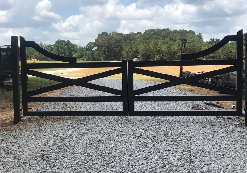 The Farmhouse Gate closed across a fresh gravel drive, two black X-braced leaves with scrolled top corners, open fields and a treeline behind.