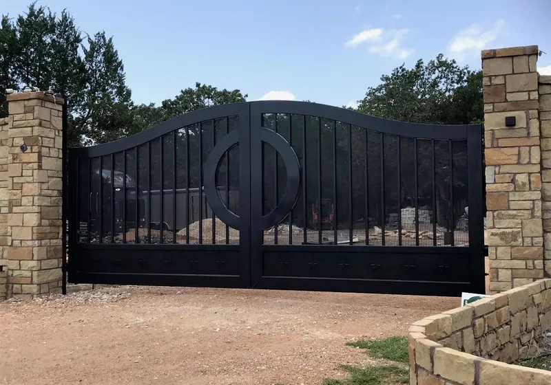 The Austin Gate closed square-on between rough stone columns on a gravel drive, its black leaves rising to a shallow ogee crest with a ring medallion at the centre and trees against a blue sky behind.