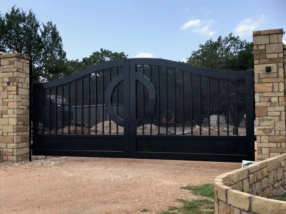 The Austin Gate closed square-on between rough stone columns on a gravel drive, its black leaves rising to a shallow ogee crest with a ring medallion at the centre and trees against a blue sky behind.