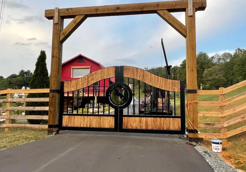 The Cypress Gate closed at the end of an asphalt drive beneath a heavy timber entrance frame, its wave-topped wood panels and black frame set against a red barn and a run of post-and-rail fence.