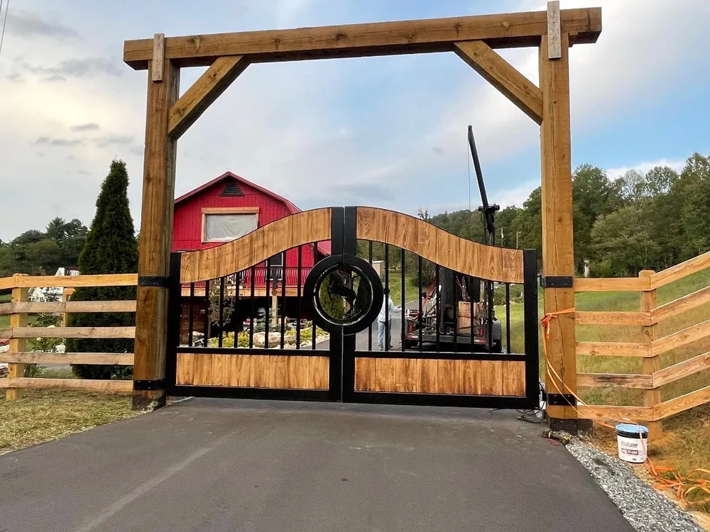 The Cypress Gate closed at the end of an asphalt drive beneath a heavy timber entrance frame, its wave-topped wood panels and black frame set against a red barn and a run of post-and-rail fence.
