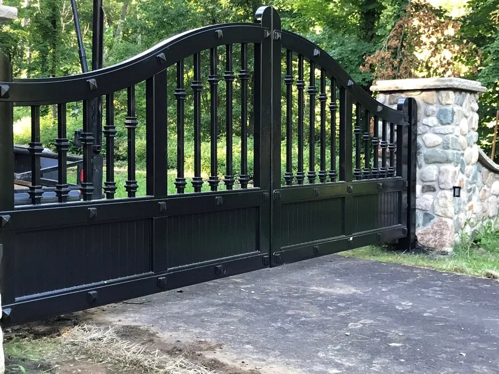 The Ella Grace Gates closed across a wooded drive, black iron balusters above a boarded lower panel, hung beside a rounded fieldstone column.