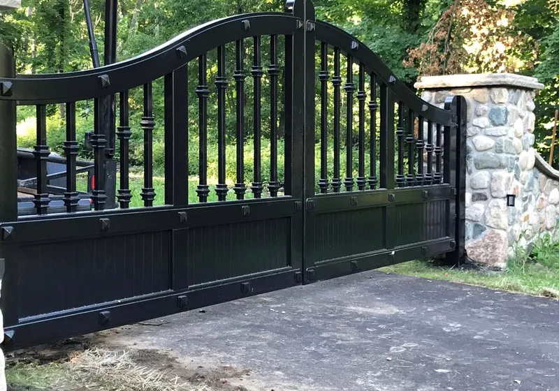 The Ella Grace Gates closed across a wooded drive, black iron balusters above a boarded lower panel, hung beside a rounded fieldstone column.