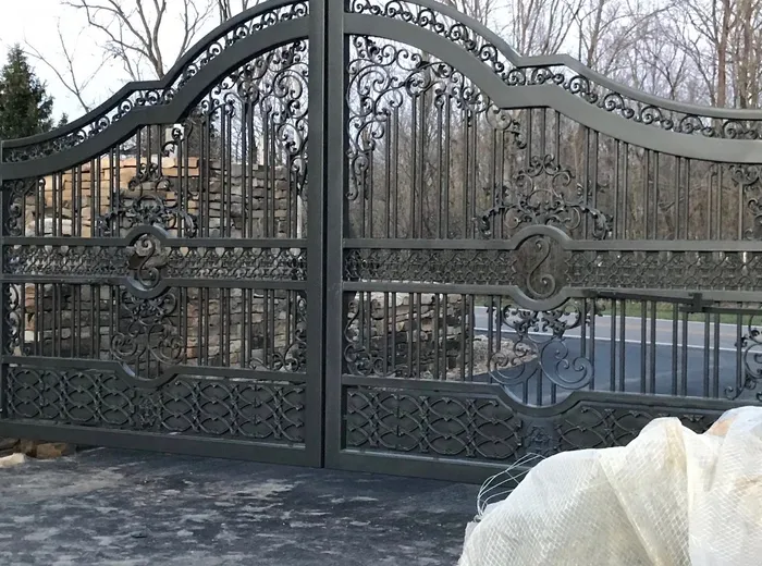 Close view of the closed leaves, showing the scrollwork crown, the rosette mid-belt and the oval medallion on each leaf, with a part-built stone wall behind the ironwork.