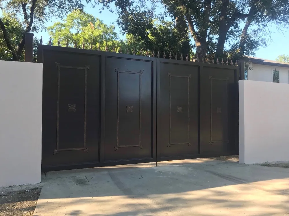 The Dallas Estate Gate closed across a concrete drive between two white stucco walls, its four dark panels topped by a row of spear finials, with oaks overhead and a pale house roof beyond the wall.