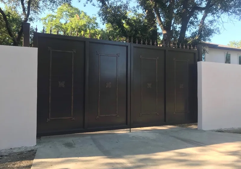 The Dallas Estate Gate closed across a concrete drive between two white stucco walls, its four dark panels topped by a row of spear finials, with oaks overhead and a pale house roof beyond the wall.