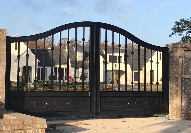 The Fresina Gate closed between brick piers at the end of a drive, its arched black leaves of collared balusters standing against a white gabled house and evening sky.