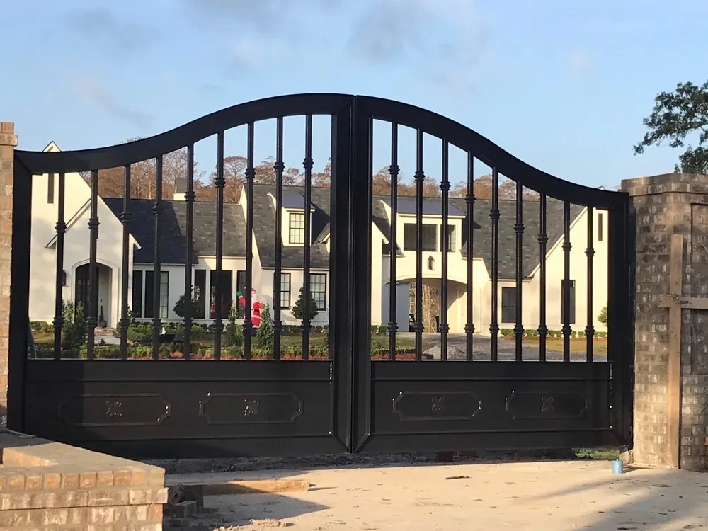 The Fresina Gate closed between brick piers at the end of a drive, its arched black leaves of collared balusters standing against a white gabled house and evening sky.