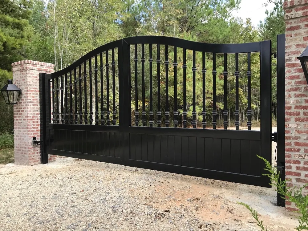 The Kling Gate closed across a gravel drive between red brick pillars, its arched top rail and collared balusters black against a stand of pines, a lantern mounted on the left pillar.