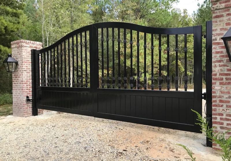 The Kling Gate closed across a gravel drive between red brick pillars, its arched top rail and collared balusters black against a stand of pines, a lantern mounted on the left pillar.