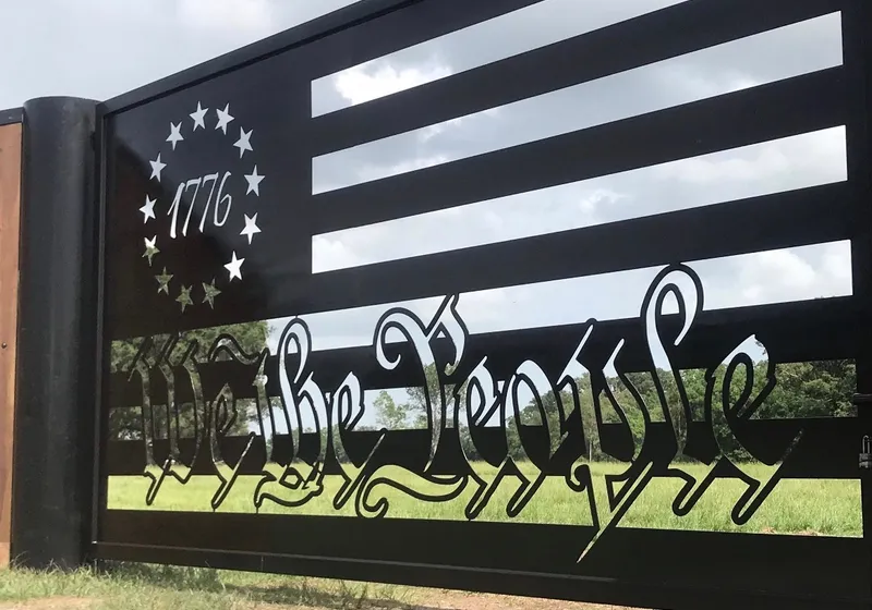 A sunlit view along The Patriot Gate, the cut star ring and the date 1776 at the left, "We the People" in blackletter across the lower stripes, and green pasture and cloud showing through the open slots.