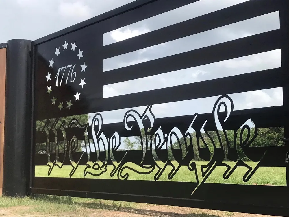 A sunlit view along The Patriot Gate, the cut star ring and the date 1776 at the left, "We the People" in blackletter across the lower stripes, and green pasture and cloud showing through the open slots.