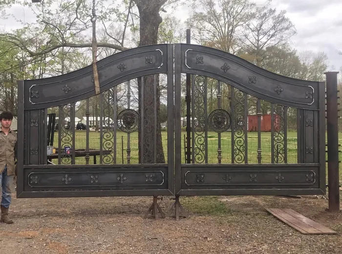 The New Yorker Gate's two leaves standing side by side on steel stands in a gravel yard, seen square-on so the curved top bands meet at the centre, the pair in a dark pewter surface with the raised mouldings and rosettes picked out lighter, and a man standing at the far left of the frame.