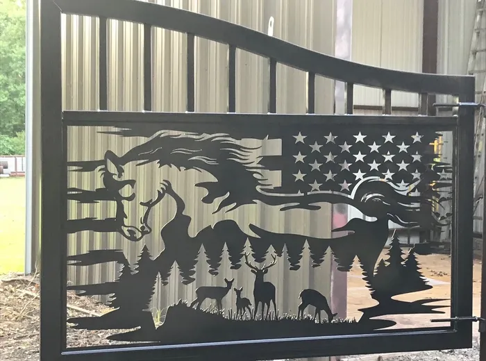 A single leaf of the Freedom Ranch Gate standing square-on in the shop against a metal building wall, the horse, flag, pines and deer all reading clearly in silhouette.