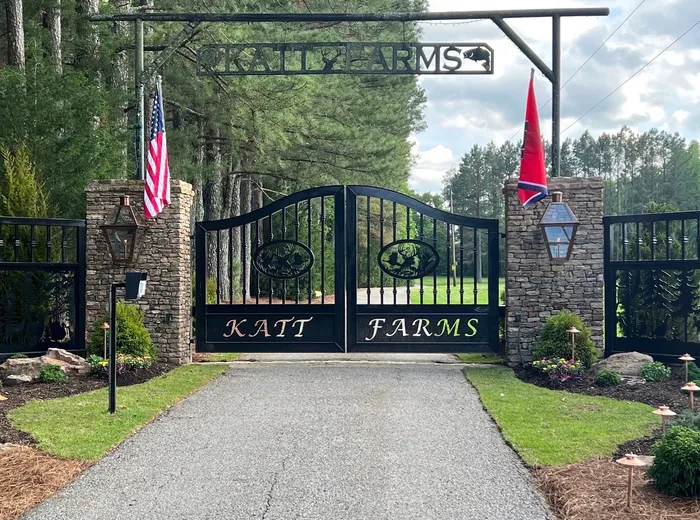 The full entrance from the road, the closed leaves between stone piers with a cut-steel name sign spanning the opening overhead on pipe posts and pines rising behind.