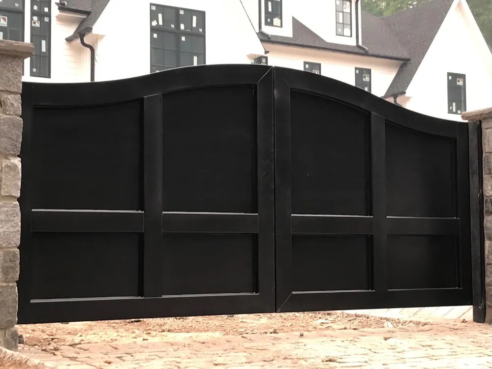 The Tappan Gate shut between rough stone piers on a cobbled drive, its black plate leaves rising to a crest at the centre, with a white gabled house behind.