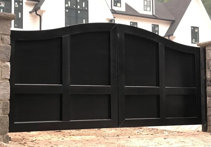 The Tappan Gate shut between rough stone piers on a cobbled drive, its black plate leaves rising to a crest at the centre, with a white gabled house behind.