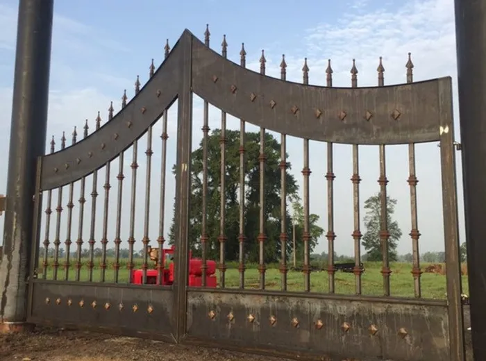 The gate in bare mill steel before the finish coat, its arch and spear finials against open sky beside one of the pipe posts.